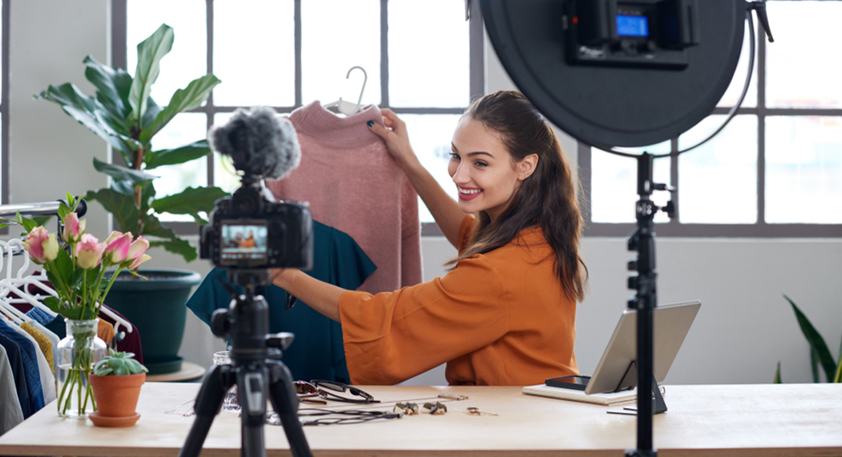 Smiling woman in an orange blouse holding a pink sweater in front of a camera while filming a fashion video at a table with clothes and plants