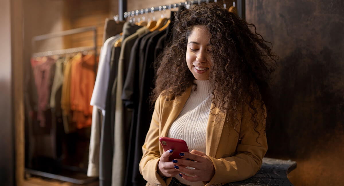 Woman with curly hair wearing a beige jacket, smiling while looking at her phone in a clothing store with racks of clothes in the background