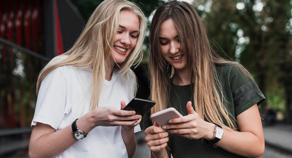 Two young women smiling and looking at their smartphones, enjoying outdoor social interaction and mobile connectivity