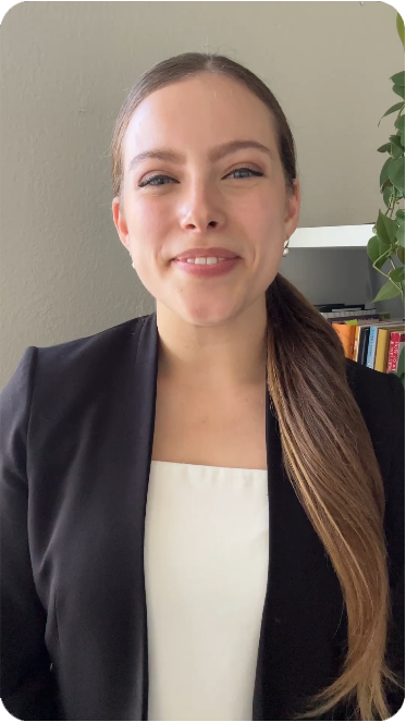 A woman with long brown hair in a ponytail, dressed in a black blazer over a white top, smiles warmly at the camera, embodying professional style